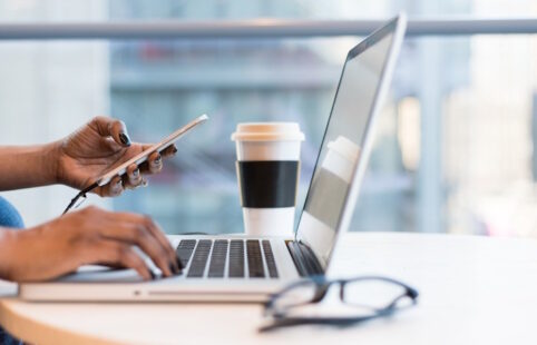 Frauenhand hält Handy vor Laptop und Kaffeebecher auf cleanem Tisch mit Brille