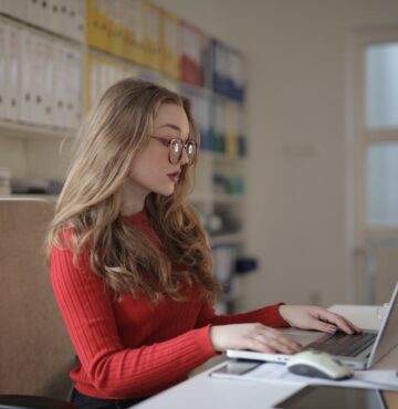 Frau in rotem Pullover mit roten großen Brillen und langem lockigem Haar sitzt am Laptop am Schreibtisch vor einer Wand mit Ordnern und Fenster