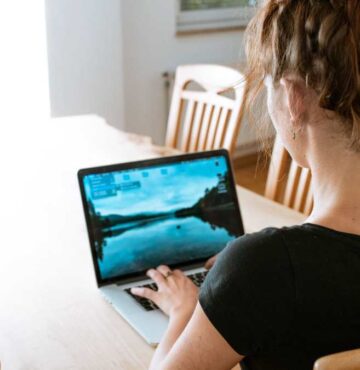 Blick über die Schulter einer Frau, die an einem Wohnzimmertisch vor einem Laptop sitzt