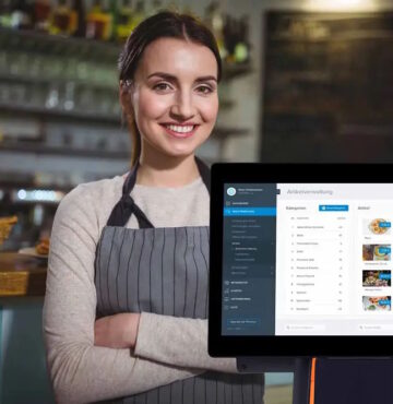 Sympathisch wirkende, lächelnde Frau, die in Café vor einer Registrierkasse mit verschränkten Armen steht.