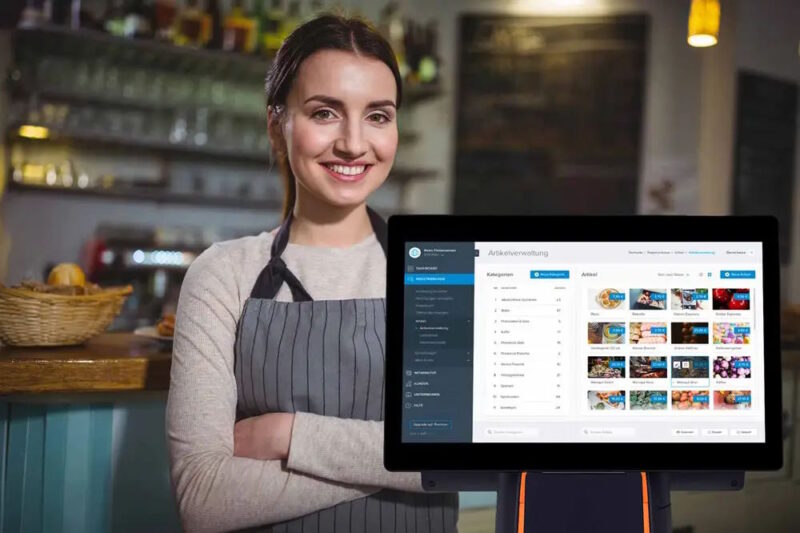 Sympathisch wirkende, lächelnde Frau, die in Café vor einer Registrierkasse mit verschränkten Armen steht.