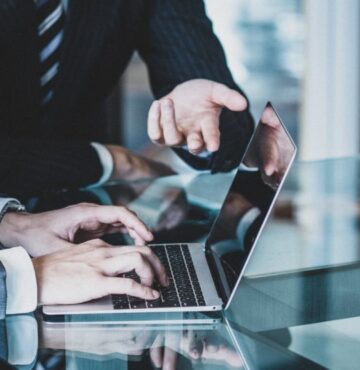 Ausschnitt von Armen zweier Männer und Schreibtischsituation, ein Mann tippt am Laptop.