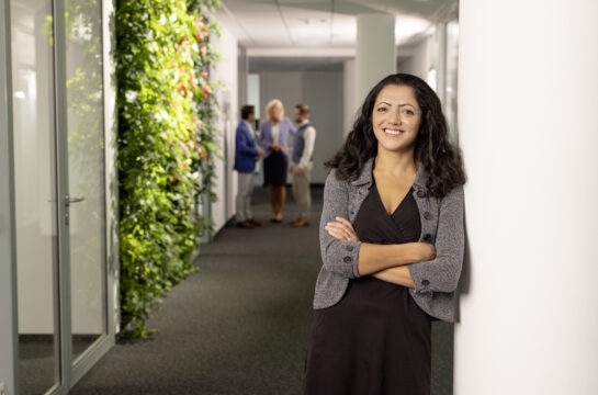 Steuerberaterin Dudu Yurttas lächelt neben der begrünten Wand in der Kanzlei in die Kamera vor drei Kollegen, die im Hintergrund stehend etwas besprechen.